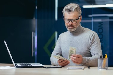 Serious mature investor working inside office, senior gray haired businessman counting money cash dollars, businessman working inside contemporary office using laptop at work.