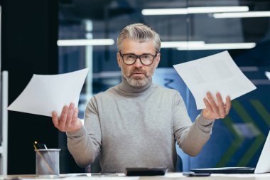 Belgelerle hayal kırıklığına uğramış ve kızgın iş adamı, finansal raporları kontrol eden olgun gri işadamı, patron başarı sonuçlarından memnun değil ve ofis içinde dizüstü bilgisayarla çalışan gelir..