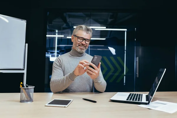 Mature successful businessman working inside office, senior gray-haired businessman using phone contentedly reading online message, man working with laptop at workplace, happy investor.