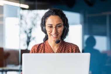 Smiling hispanic businesswoman working inside office with laptop and headset for video call, woman sitting at workplace happy working with clients.