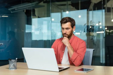 Serious thinking businessman close up, man in red casual shirt working inside modern office, mature boss working with laptop at workplace while sitting