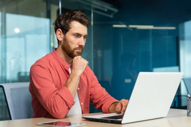 Serious thinking businessman close up, man in red casual shirt working inside modern office, mature boss working with laptop at workplace while sitting