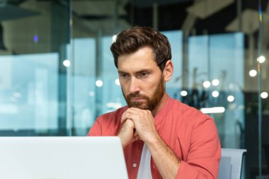 Serious thinking businessman close up, man in red casual shirt working inside modern office, mature boss working with laptop at workplace while sitting