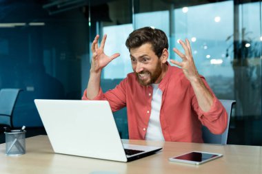 Angry and nervous businessman talking on video call remotely, man shouting to colleagues online, boss in shirt working inside modern office using laptop at work.