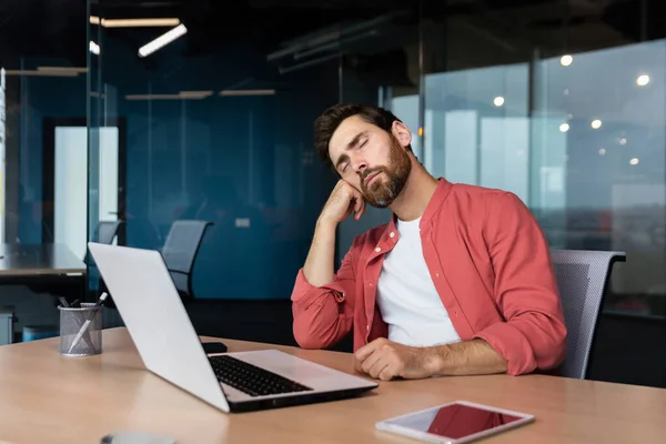 Tired businessman in casual red shirt sleeps at workplace, man on desk fell asleep during working hours inside office with laptop.