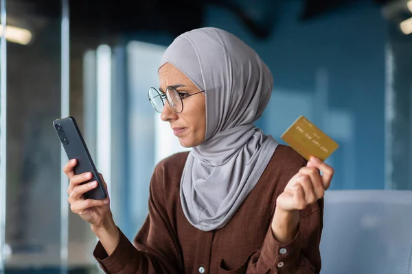 Close-up photo. A young Arab woman in a hijab and glasses is holding a phone and a credit card. Online shopping, payment, checking account.