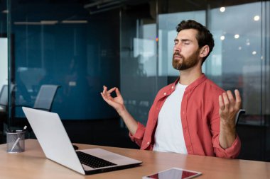 Businessman in a shirt resting and meditating in the office in the lotus position, a man at the workplace relaxes during the day sitting at the workplace with a laptop with his eyes closed.