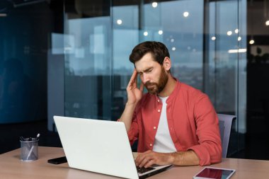 Sick man at workplace, mature businessman has severe headache, boss in shirt works inside office with laptop.