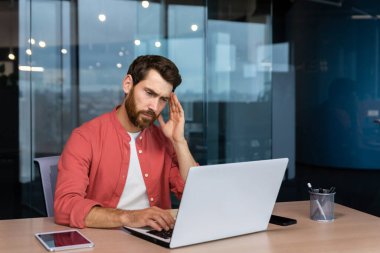 Sick man at workplace, mature businessman has severe headache, boss in shirt works inside office with laptop.