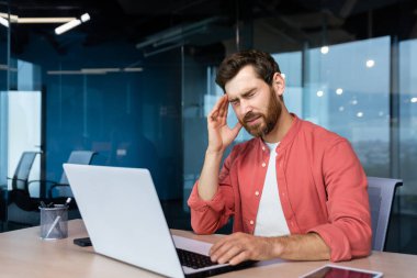 Sick man at workplace, mature businessman has severe headache, boss in shirt works inside office with laptop.