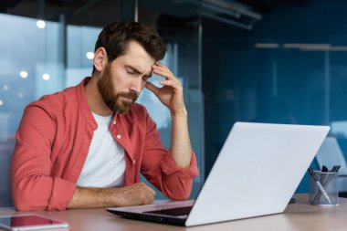 Frustrated businessman depressed at workplace working on laptop, man in shirt upset and sad displeased with bad work results and achievement inside office.
