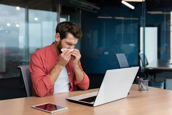 Sick man at work has flu and cold, businessman sneezes and coughs at workplace working inside office at desk using laptop at work.