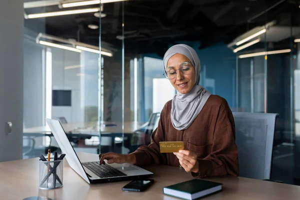A young Muslim woman in a hijab is sitting in the office at a table with a laptop, holding and using a credit card. Pays bills, checks, online shopping.