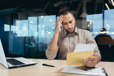 An upset young African-American man reads bad news from a letter he received. He is sitting at the desk in the office, holding his head with his hand.