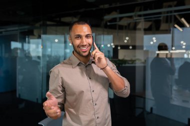 A young African American man is standing in the office and talking on the phone. gesturing with his hands, looking at the camera, smiling.