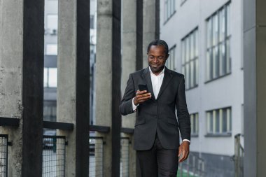 A successful mature African American man in a business suit is walking outside an office building, using a phone, typing a message and looking at the news online while smiling
