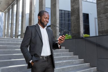 A successful mature African American man in a business suit is walking outside an office building, using a phone, typing a message and looking at the news online while smiling