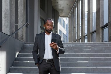 Serious and thoughtful businessman reading online news on phone african american boss in suit outside office building looking at smartphone screen concentration, walking near building in background.