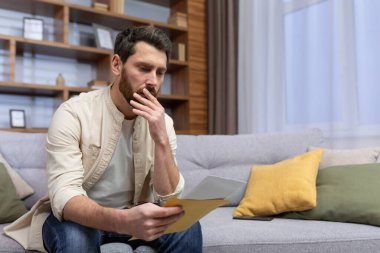 Shocked young man sitting on the sofa at home, holding a letter in his hands. Got bad news, divorce papers, financial debt, medical results.