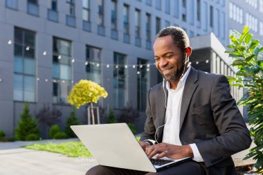 Businessman, African American freelancer sitting near office center, orking online on laptop and wearing headphones. He types on the keyboard, communicates through a video call, smiles.