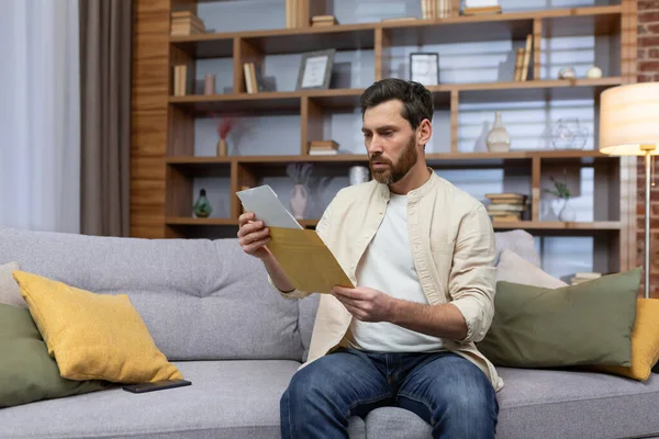Upset young man sitting on sofa at home, received a letter, reads bad news. Divorce documents, financial debt, rent, credit, Upset young man sitting on sofa at home, received a letter, reads bad news