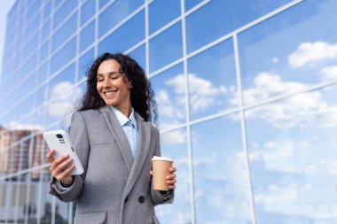 Portrait of a young beautiful Latin American businesswoman in a suit walking outside near a glass office center. Holds a cup of coffee, looks at the phone, smiles.