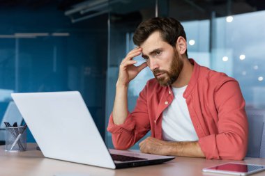 A stressed and tired young businessman, student, freelancer in a red shirt sits in the office at a laptop. holding his head. Overtime work, deadline, problems.