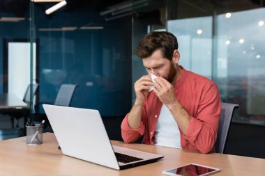 A young man is sick at work. sits at the desk in the office, wipes his nose with a napkin, has a runny nose, does not feel well.