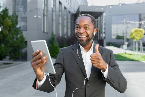 A young African American man is standing outside near an office center wearing headphones and talking on a video call. Holds a tablet in his hands, shows a super, smiles, looks at the camera.