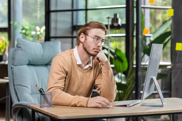 Frustrated and upset businessman reading news online from computer monitor, young blond man sad at workplace inside office in casual clothes unhappy with results.