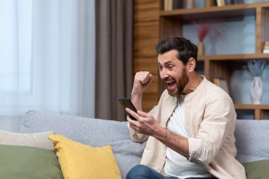 A handsome young man in a beige shirt is sitting on the sofa at home and playing games on the phone. Received good news, messages, rejoices, celebrates, shouts, shows a yes gesture with his hand.