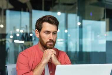Close-up photo. Concentrated and thoughtful young man in a red shirt works, studies on a laptop in the office