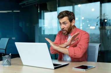 Problems at work. An angry young man in a red shirt is sitting in the office and talking on a video call from a laptop. Screams, aggressively gestures with his hands, denies.