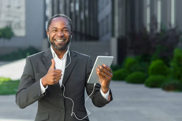 A young African American man in a suit is standing outside near an office center wearing headphones, holding a tablet. Looks into the camera. smiles, points with a super finger.