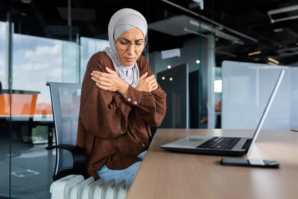 Freezing arab woman inside office, business woman in hijab cold indoor heating and heating not working, using laptop in work at workplace, sitting trying to warm.