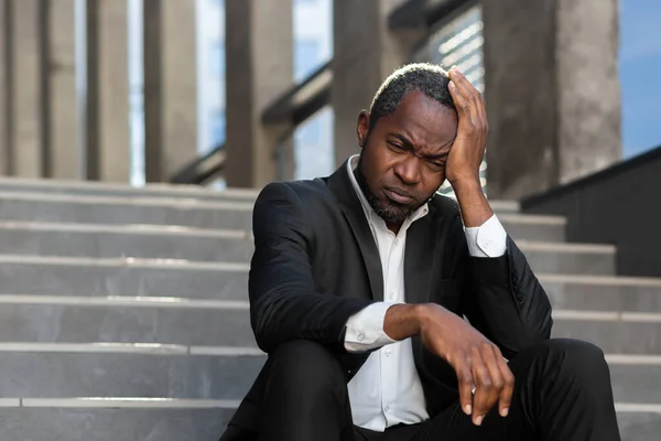 Sad and upset African-American man in a suit sits on the stairs near the office center, looks thoughtfully down, holds his head in his hand.