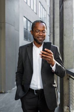 Dikey fotoğraf. Ofis merkezinin yanında elini cebinde tutan takım elbiseli ve gözlüklü gülümseyen Afrikalı Amerikalı bir işadamının portresi. Telefonu kullanıyor, arama yapıyor, yazılar yazıyor.