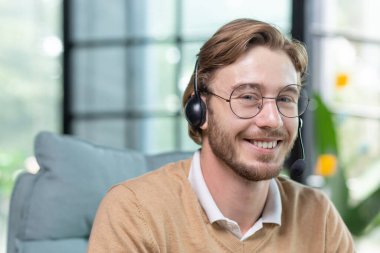 Video görüşmesi için kulaklığı olan genç bir adamın portresi, gözlüklü yakın çekim teknisyen gülümseyen ve kameraya bakan, iş adamı sarışın müşteri hizmetleri online.