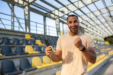 Eğitimin sonuçlarından memnun olan başarılı bir adam, kameraya bakar ve gülümser, İspanyol elinde bir telefonla online bir spor programı kullanır, bir atlet başarıyla elini kaldırır..