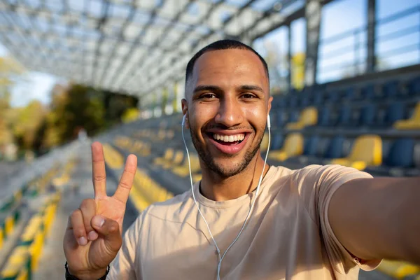 Sportsman runner in stadium taking selfie photo and talking on video call with friends, hispanic man looking at camera and smiling recording sports blog, young man after active training.