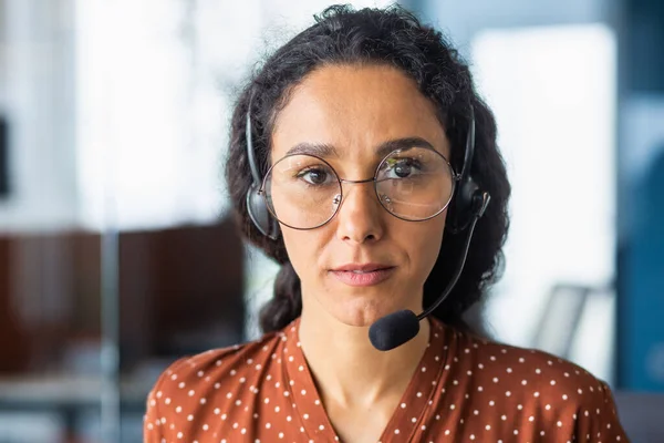 Serious and concentrated woman with video call headset looking at camera, hispanic woman thinking close up inside office, call center and support tech worker in glasses.
