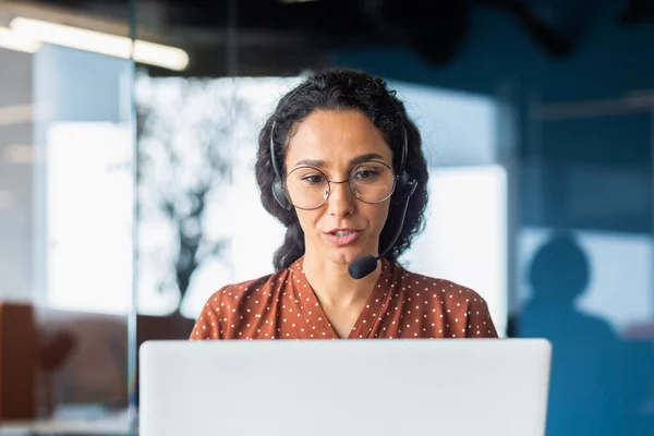 Hispanic woman with online meeting video call headset and laptop talking to client remotely, woman in glasses and curly hair inside office, support helpline worker.