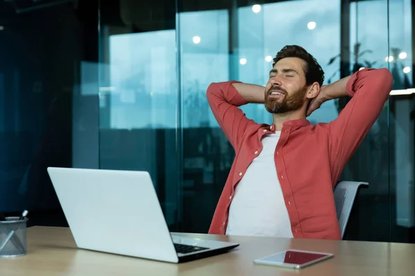 Successful businessman in office in a red shirt rests with his hands folded head with closed eyes, mature man with a beard works with a laptop at the workplace inside, dreams of future achievements.