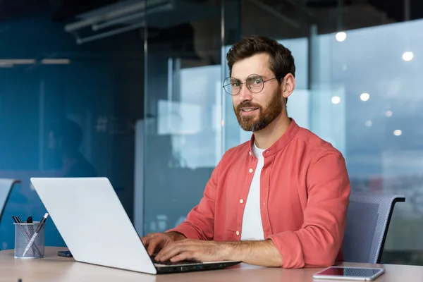 Serious and focused mature businessman in red shirt wearing glasses looking at camera, young man at work typing on laptop keyboard, programmer developer portrait at work.