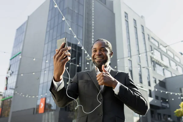 An African American businessman in a suit is standing outside an office center wearing headphones and holding a phone. Talks on a video call, points a super finger at the camera, smiles.