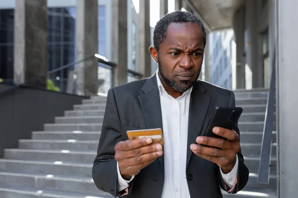 A problem with the account. Upset african American man in a suit standing outside a bank office. He is holding a credit card. He looks worriedly at the phone, dials, checks, pays.