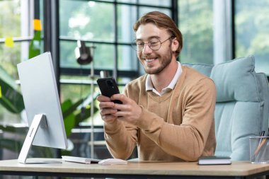 Genç yakışıklı iş adamı, serbest çalışan, bilgisayarın başında oturuyor ve telefonu kullanıyor. Mesajlar yaz, kahkahalar at, haberleri oku, gülümse.