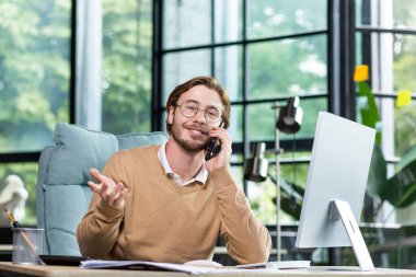 Genç yakışıklı iş adamı, ofiste bilgisayar başında oturan ve telefonda konuşan serbest çalışan. Kameraya bakıyor. Jest, gülümseme.