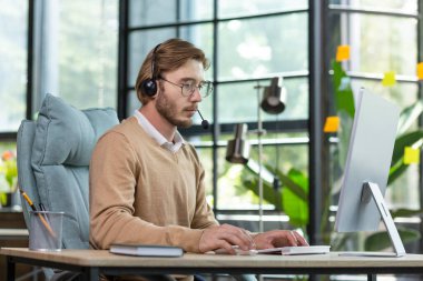 Çevrimiçi ticaret, destek, danışma. Masada, kulaklıkla oturan genç yakışıklı bir adamın portresi, bilgisayar başında çalışmaya odaklanıyor..