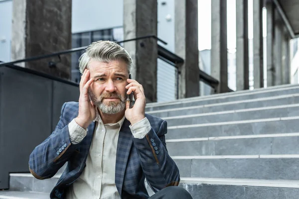 Upset and disappointed mature businessman sitting on stairs of office building outside, man in business suit bankrupt talking on the phone, senior boss investor lost money
